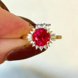May include: A gold ring with a large, round, bright red gemstone encircled by smaller white stones and baguette-cut stones. The ring is held up by a hand with a dark brown manicure. The text "DiamondsPage" is visible.