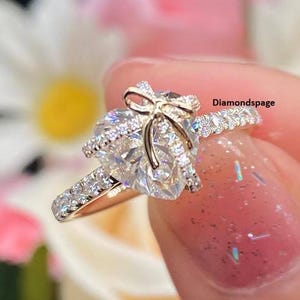 May include: A gold ring with a pear-shaped diamond center stone, accented with a bow design and smaller diamonds along the band. The ring is held up against a blurred floral background. The text "Diamondspage" is visible.