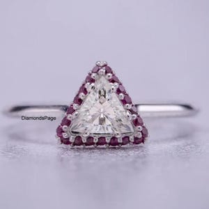 May include: A silver ring featuring a triangular, white gemstone center, surrounded by a halo of small, red gemstones. The ring's band is thin and polished, set against a soft, blurred background.