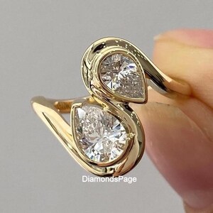 May include: A bypass ring crafted from polished yellow gold featuring two pear-cut diamonds set in a smooth bezel setting. The design creates an S-shaped silhouette with the stones positioned at opposite ends. The text DiamondsPage is visible.