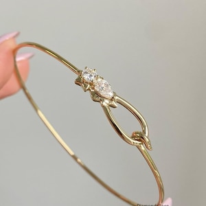 Könnte beinhalten: Ein filigranes goldenes Armreif-Armband mit einem Knotendesign und zwei klaren Edelsteinen. Das Armband hat ein schlankes, poliertes Band und ein dekoratives Mittelstück mit einem runden und einem birnenförmigen Stein.