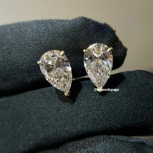 Może przedstawiać: Para kolczyków z diamentowymi wstawkami w kształcie gruszki, osadzonymi w złotej oprawie. Diamenty są przejrzyste i błyszczące.