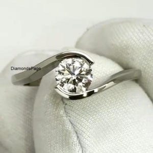 May include: A silver engagement ring featuring a round, clear diamond in a bezel setting. The band curves around the diamond, creating a modern design. The ring is displayed against a white background.