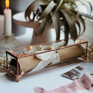 May include: A rectangular glass display box with a copper-toned metal frame and ornate corner accents. Inside, a rolled brown paper scroll is tied with a cream-colored satin ribbon. The box features a hinged lid with a small metal latch.