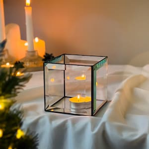 May include: A clear glass candle holder with a square shape and teal trim. Inside, a lit tea light candle casts a warm glow. Other lit candles are in the background, creating a cozy ambiance. The holder sits on a white surface.