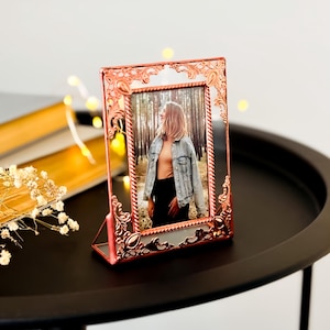 May include: A decorative, rose-gold colored photo frame with an ornate, filigree design. The frame holds a photograph of a person in a forest setting. The frame is displayed on a black table, with soft, out-of-focus lights in the background.