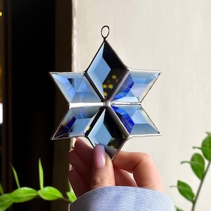 Puede incluir: Un adorno de estrella de vidrio azul y plateado con un diseño geométrico. La estrella cuelga de un lazo plateado.