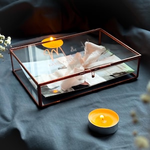 May include: A clear glass box with a copper frame and lid. The box is decorated with the initials "M & A" and the words "Melanie & Andrew" in a script font. The box is sitting on a blue surface with two lit candles.
