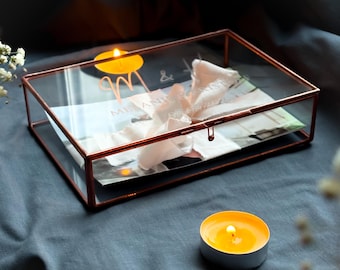 May include: A clear glass box with a copper frame and lid. The box is decorated with the initials "M & A" and the words "Melanie & Andrew" in a script font. The box is sitting on a blue surface with two lit candles.