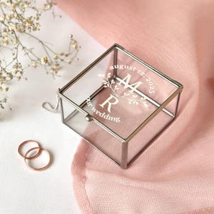 Könnte beinhalten: Eine transparente Glas-Ringbox mit silbernen Rändern, graviert mit den Initialen "M R" und dem Datum "12. August 2025". Zwei goldene Eheringe liegen neben der Box auf einem rosa Stoff.