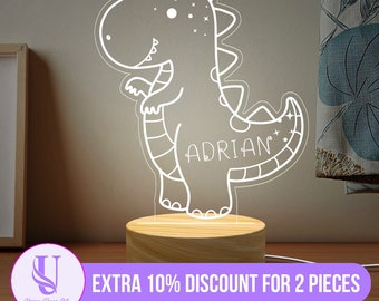 Puede incluir: Una luz nocturna de dinosaurio blanco con el nombre "ADRIAN" en el lateral, sentada sobre una base de madera. La luz está encendida e ilumina el dinosaurio.