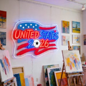 May include: A neon sign in the shape of the American flag, with the words "UNITED STATES 2026" in red and blue. A soccer ball is in the center. The sign is in an art studio with paintings on easels and the walls.