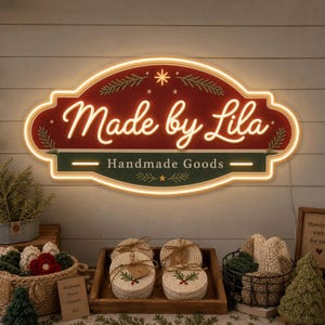 May include: A horizontal, oval-shaped LED neon sign with a red and dark green background. The sign features the white text "Made by Lila" in a script font and "Handmade Goods" in a sans-serif font, accented by white pine branch graphics.
