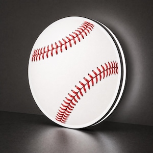 May include: A circular wall light fixture shaped like a baseball. The surface is white with the characteristic red stitching pattern of a baseball. The light emits a soft glow from the edges, creating a halo effect around the circular frame.