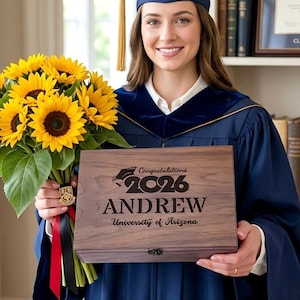 Op de afbeelding: Een houten afstudeergeschenkdoos, gegraveerd met "Congratulations 2026 ANDREW University of Arizona." De doos wordt vastgehouden door een persoon in een afstudeer toga en hoed, met een boeket zonnebloemen.