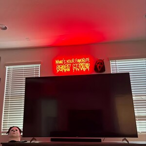 Peut inclure: Un t&eacute;l&eacute;viseur &agrave; &eacute;cran plat noir est mont&eacute; sur un mur avec une enseigne au n&eacute;on rouge qui dit "Quel est votre film d'horreur pr&eacute;f&eacute;r&eacute; ?" Un masque de fant&ocirc;me rouge et blanc est sur le mur &agrave; droite de l'enseigne.