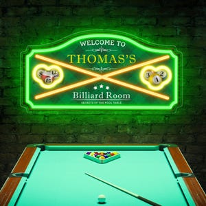 Può includere: Un'insegna al neon con la scritta "WELCOME TO THOMAS'S BILLIARD ROOM" è esposta sopra un tavolo da biliardo. L'insegna ha un bordo verde e presenta linee gialle incrociate con le palle da biliardo. Il tavolo da biliardo ha una superficie in feltro verde.