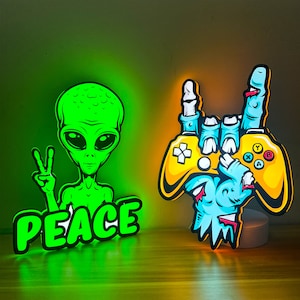 May include: Two neon signs, one with a green alien holding a peace sign and the word "PEACE" below, and the other with a blue hand holding a yellow video game controller with the words "A", "B", "X", and "Y" on it.
