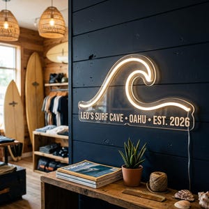 Puede incluir: Un letrero de neón personalizado con forma de ola oceánica estilizada montado en una pared de tablones de madera azul oscuro. El letrero presenta el texto blanco LEO'S SURF CAVE • OAHU • EST. 2026 y emite un brillo blanco cálido desde el tubo.