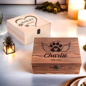 Puede incluir: Dos cajas de recuerdos de madera. Una es de color claro con un diseño de corazón y huella de pata, el nombre "Bailey" y fechas. La otra es de color marrón con una huella de pata, alas, el nombre "Charlie" y fechas. Ambas tienen cierres metálicos.