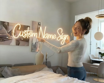 May include: A neon sign that reads "Custom Name Sign" in a cursive font. The sign is illuminated with a warm, soft glow. The sign is held up by a person in a white sweater and blue jeans. The background includes a bed, artwork, and decorative lighting.