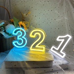 Könnte beinhalten: Drei Neon-Lichtzahlen, 3, 2 und 1, in blau, gelb und weiß, jeweils. Die Zahlen befinden sich auf einer weißen Oberfläche mit weißem Hintergrund.