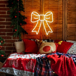 May include: A white neon bow sign with a warm glow, hanging on a wooden wall above a bed with red and white bedding. The bed is decorated with red and white pillows, including a reindeer pillow with a red nose.
