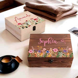 May include: Two wooden keepsake boxes, one light-colored and one dark-colored, with floral designs. The light box has the name "Amanda" in red script. The dark box has the name "Sophia" in pink script. Both have metal clasps.