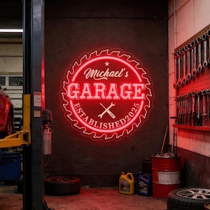 Peut inclure: Enseigne au néon rouge avec l'inscription "Michael's GARAGE ESTABLISHED 2025" dans un design circulaire, avec un motif de clé et de tournevis. L'enseigne est fixée sur un mur sombre dans un garage, avec des outils et équipements visibles.