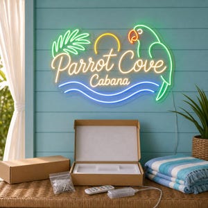Könnte beinhalten: Ein Neonschild mit dem Text Parrot Cove Cabana in Gelb, einem grünen Papagei, einer gelben Sonne und blauen Wellenlinien. Das Schild ist an einer hellblauen Wand über einem offenen Karton, einer Fernbedienung und einem blau gestreiften Handtuch montiert.