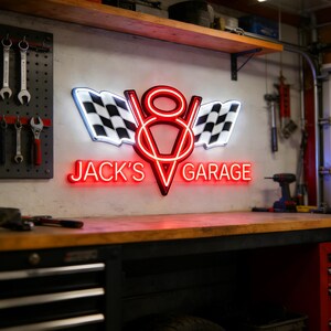 Può includere: Un'insegna al neon con un design a motore V8 rosso e bianco e bandiere a scacchi. L'insegna recita "JACK'S GARAGE" in neon rosso. È montata su un muro sopra un banco da lavoro in legno in un'officina.