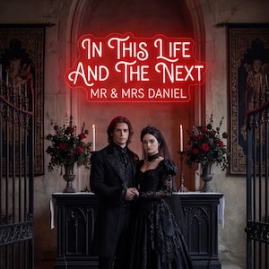 Op de afbeelding: Een stel in zwarte kleding staat voor een altaar. De vrouw draagt een zwarte jurk en sluier, de man een zwart pak. Boven hen staat in rode neonletters "In This Life And The Next" en "MR & MRS DANIEL".