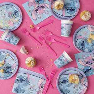 May include: A party supply set featuring Stitch and Angel from Lilo and Stitch, including light blue and purple paper plates, paper cups, napkins, and pink plastic forks, with several cupcakes scattered across the surface.
