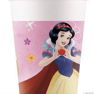 May include: A white paper cup featuring a pink band decorated with a graphic illustration of Snow White holding a red apple. The design includes small yellow and purple flowers and white star shapes against a soft pink background.
