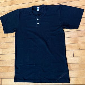 May include: A black short-sleeved Henley shirt with two white buttons. The shirt has a crew neck and a small logo tag at the neckline. The shirt is made of a soft material.