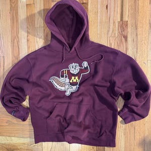 May include: A maroon hooded sweatshirt with a kangaroo pocket and drawstring. The sweatshirt features a cartoon squirrel mascot wearing a sweater with a gold "M" on the front. The mascot is on a white background. The sweatshirt is on a wooden surface.