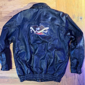 May include: Black leather jacket with a zipper closure and a ribbed hem. The back of the jacket features an embroidered emblem with an eagle, banner, and American flag colors. The jacket has a classic bomber style.
