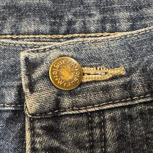 May include: Close-up of blue denim jeans with a button closure. The button is gold-colored and embossed with the word "Wrangler" in a circular design. The jeans have a classic five-pocket design.
