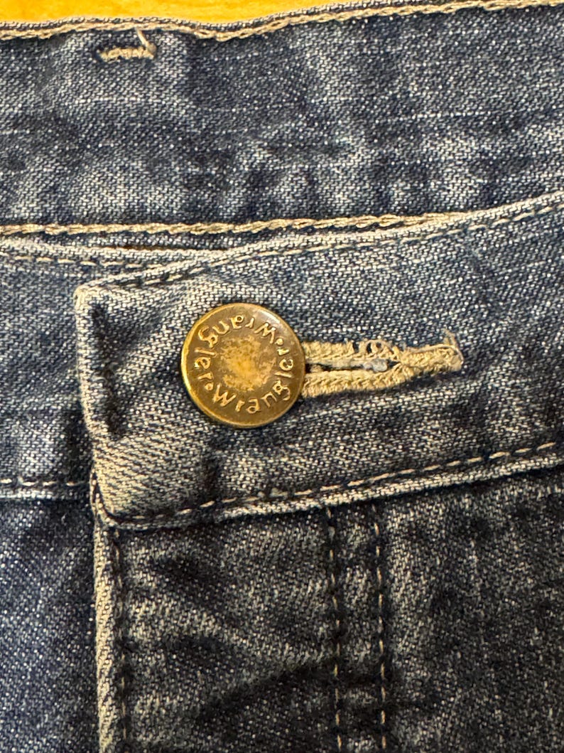 May include: Close-up of blue denim jeans with a button closure. The button is gold-colored and embossed with the word "Wrangler" in a circular design. The jeans have a classic five-pocket design.