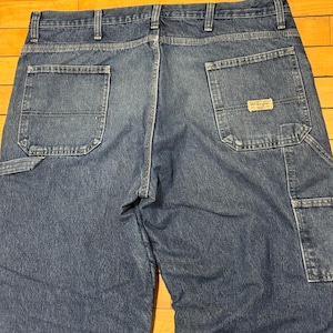 May include: Close-up of the back of blue denim work pants. The pants have two large back pockets, a small pocket, and a Wrangler brand patch. The denim is a medium wash with visible stitching and reinforced seams.