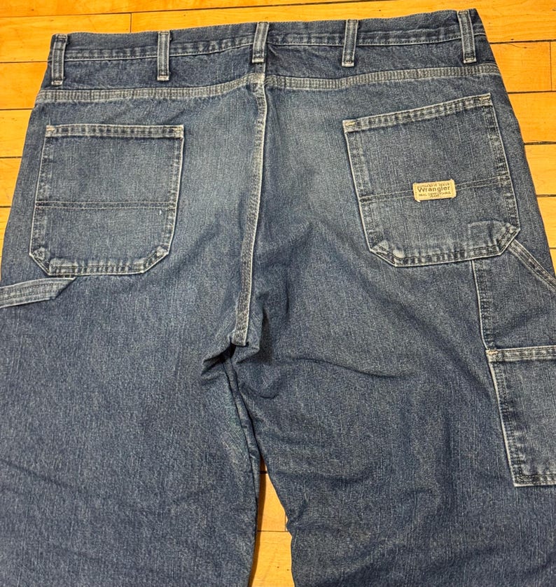 May include: Close-up of the back of blue denim work pants. The pants have two large back pockets, a small pocket, and a Wrangler brand patch. The denim is a medium wash with visible stitching and reinforced seams.