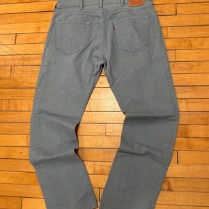 May include: Light blue denim jeans with classic five-pocket styling. Features a red Levi's tag on the back pocket. The jeans are laid flat on a wooden surface.