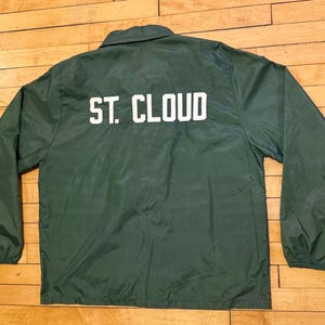 May include: A dark green coach jacket with the text "ST. CLOUD" printed in white across the back. The jacket has a collared neckline, long sleeves, and a button-up front. The jacket is made of a lightweight, possibly nylon, material.