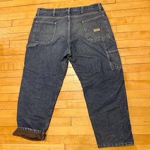 May include: Blue denim work pants with multiple pockets, including a carpenter pocket. The pants have a brown insulated lining and a small brand label on the back pocket. The jeans are laid flat on a wooden surface.