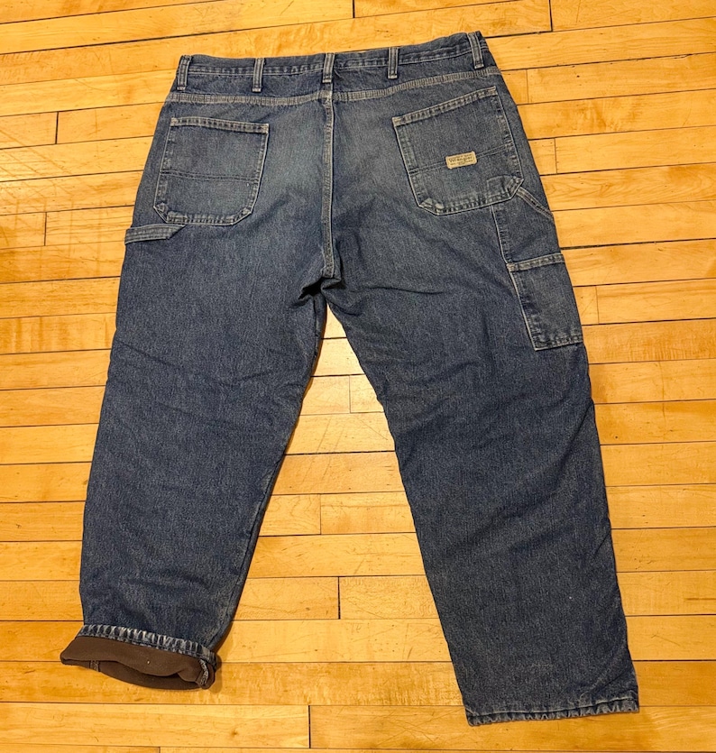 May include: Blue denim work pants with multiple pockets, including a carpenter pocket. The pants have a brown insulated lining and a small brand label on the back pocket. The jeans are laid flat on a wooden surface.
