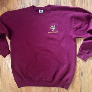 May include: A maroon Champion sweatshirt with a crew neck and long sleeves. The sweatshirt features a small graphic and the text "Minnesota Supremes" on the front. The Champion logo is visible on the upper chest.