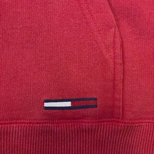 K&ouml;nnte beinhalten: Ein roter Kapuzenpullover mit einem kleinen gestickten Logo, das ein wei&szlig;es Rechteck mit einem blauen Streifen auf rotem Hintergrund zeigt.