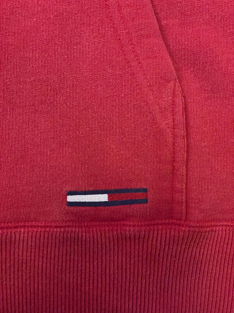 K&ouml;nnte beinhalten: Ein roter Kapuzenpullover mit einem kleinen gestickten Logo, das ein wei&szlig;es Rechteck mit einem blauen Streifen auf rotem Hintergrund zeigt.