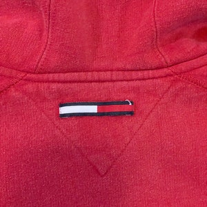 K&ouml;nnte beinhalten: Roter Kapuzenpullover mit einem kleinen rechteckigen Etikett in Wei&szlig;, Rot und Blau, das auf der R&uuml;ckseite eingen&auml;ht ist.