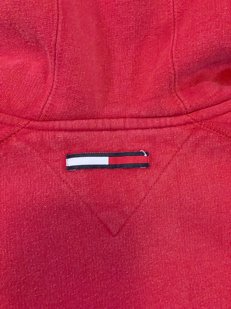 K&ouml;nnte beinhalten: Roter Kapuzenpullover mit einem kleinen rechteckigen Etikett in Wei&szlig;, Rot und Blau, das auf der R&uuml;ckseite eingen&auml;ht ist.